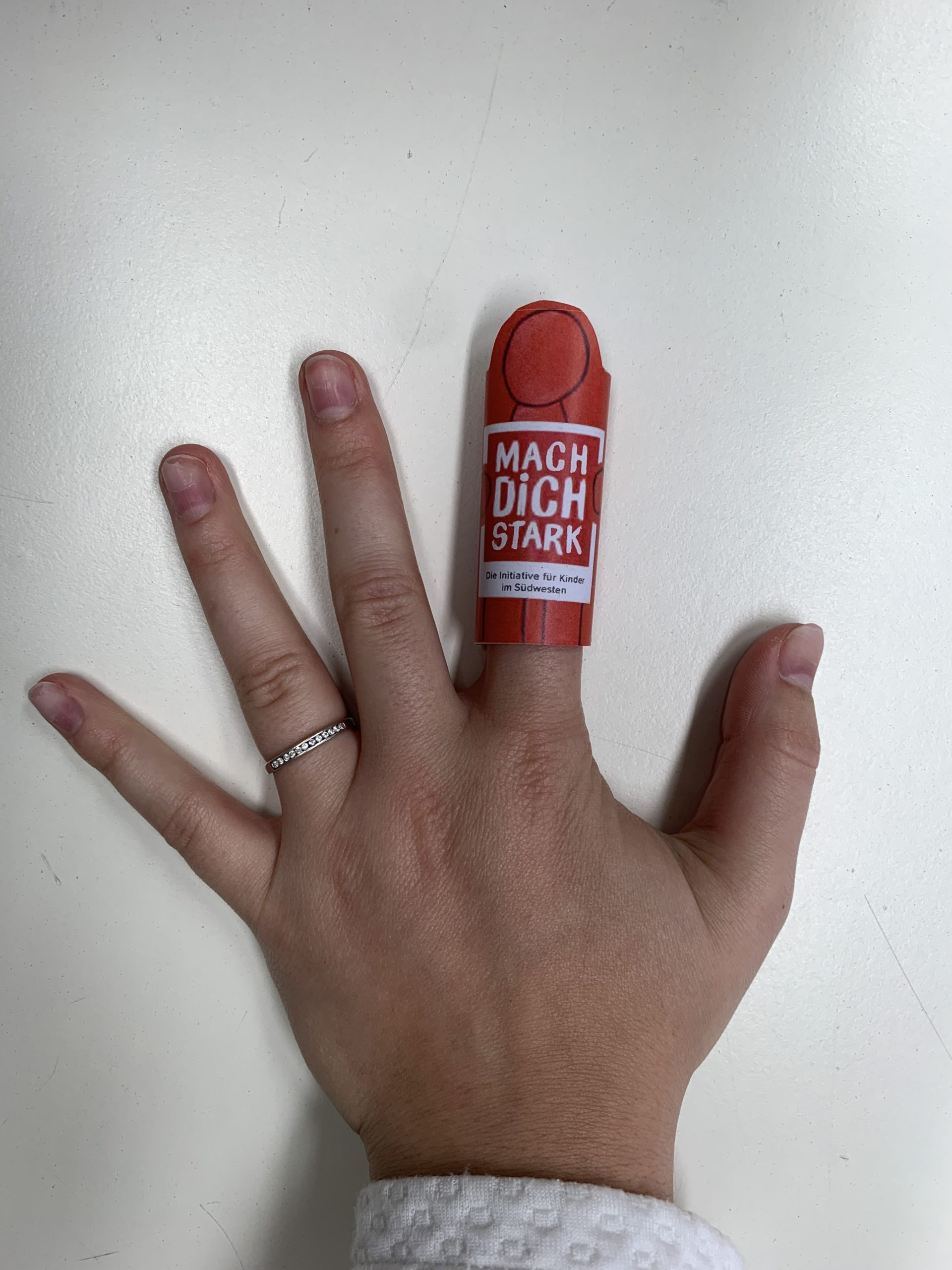 Eine Hand mit der Starkmacher-Fingerpuppe am Zeigefinger. Die Fingerpuppe ist eine rote Figur mit der Aufschrift MACH DICH STARK