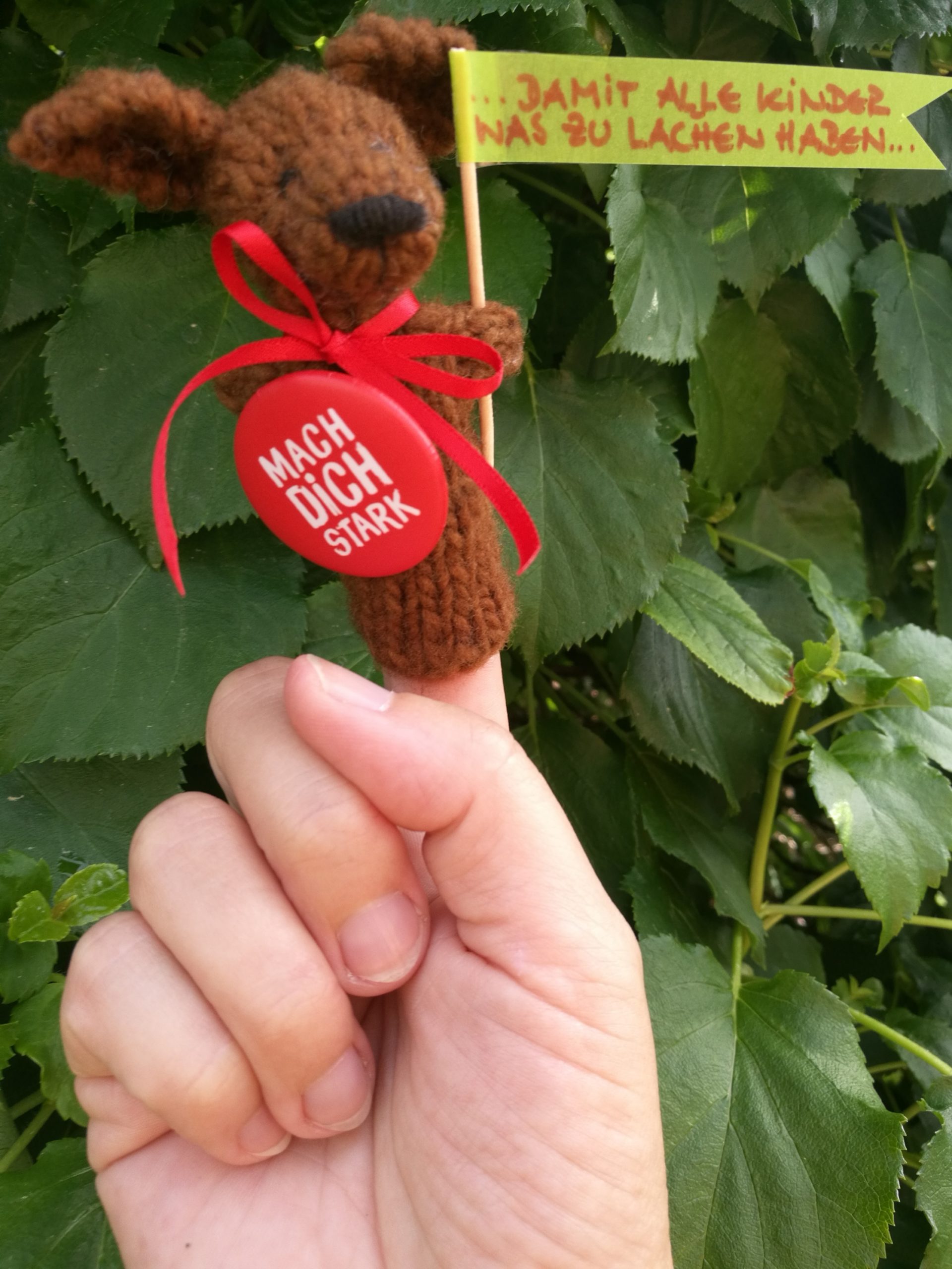 Eine Hand mit einer Fingerpuppe. Die Puppe hält eine Fahne mit der Aufschrift: "Damit alle Kinder was zu lachen haben".