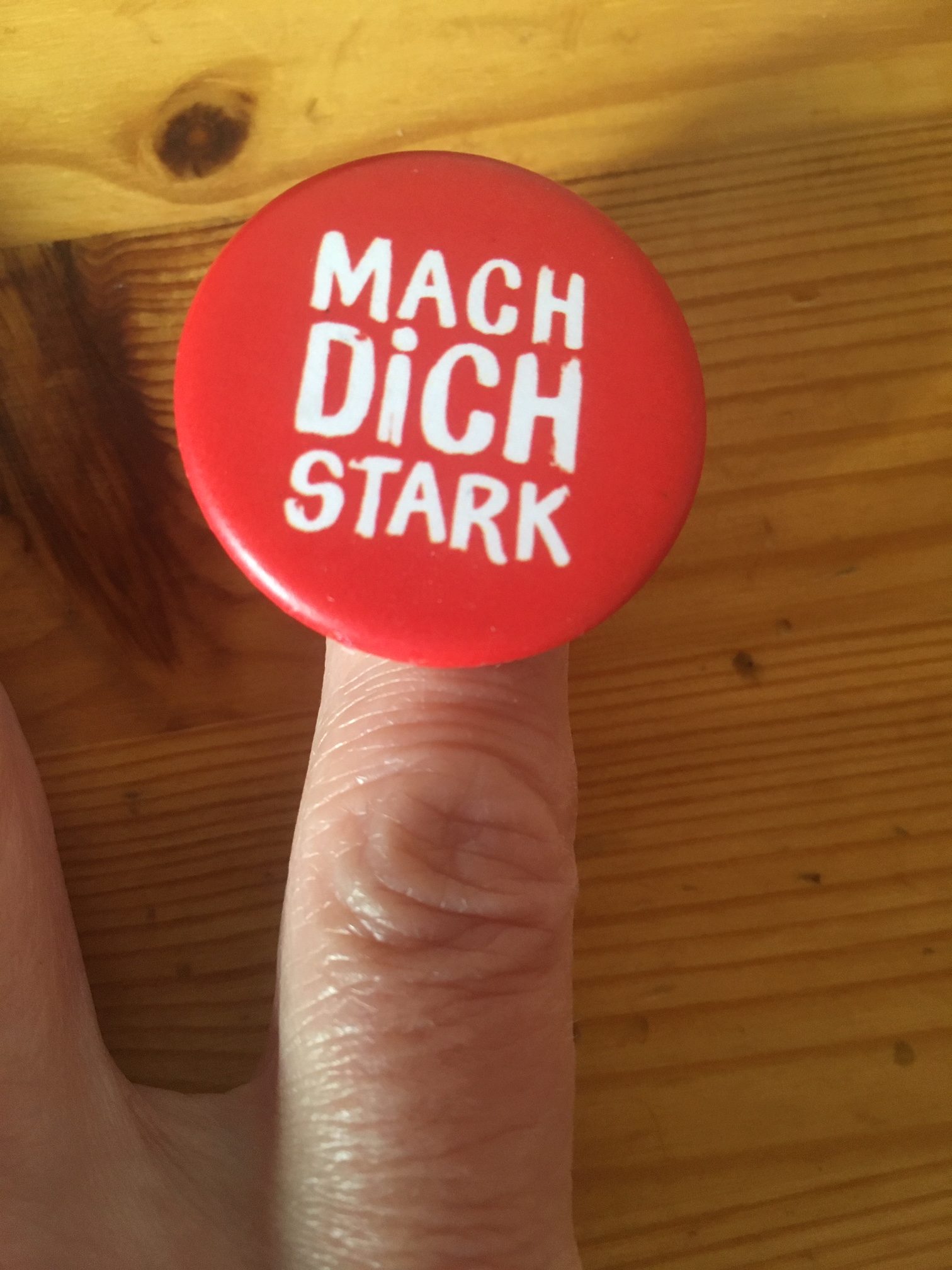 Der Zeigefinger einer Hand mit einem runden, roten Button. Im Hintergrund ein Holztisch.