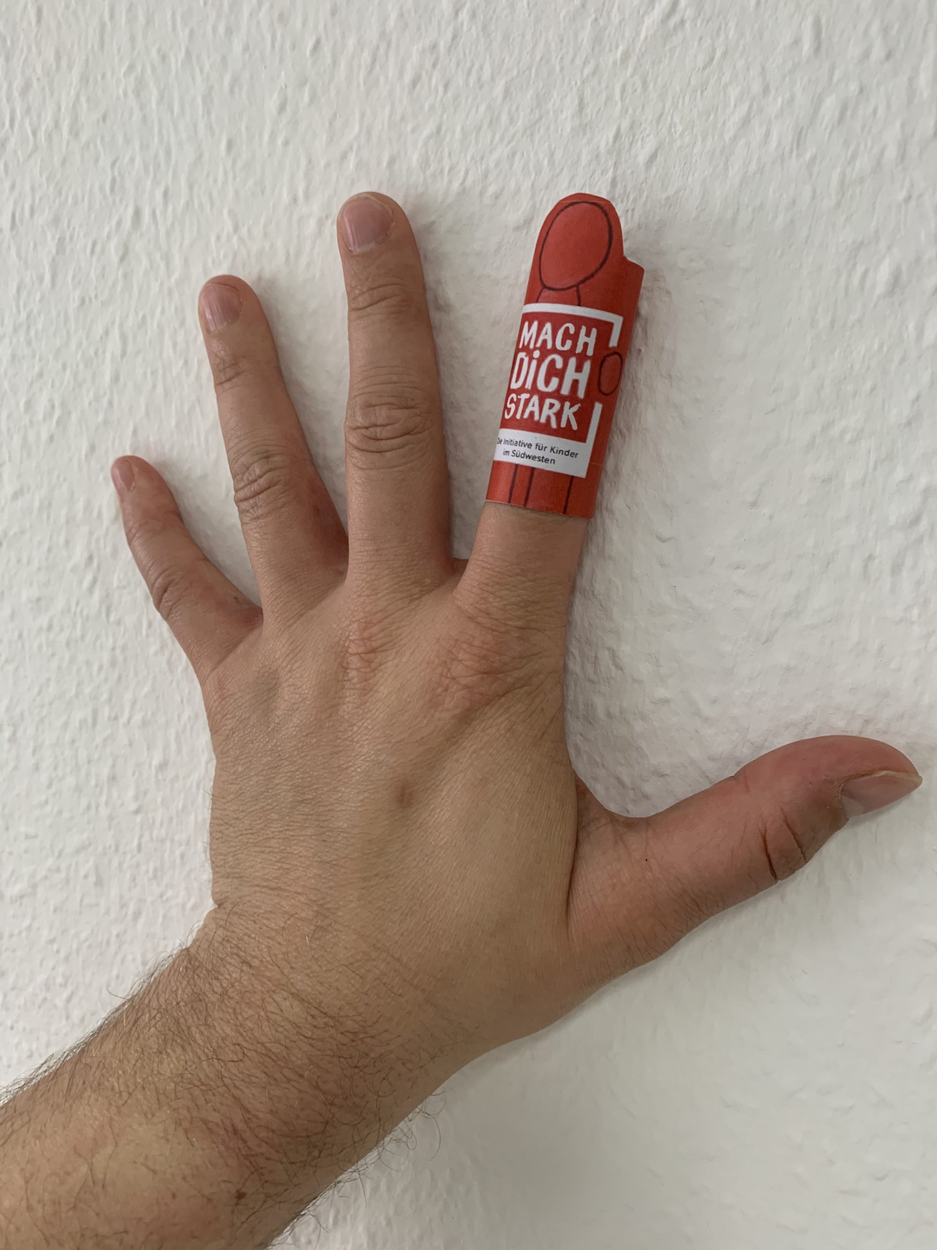 Eine Hand mit der Starkmacher-Fingerpuppe am Zeigefinger. Die Fingerpuppe ist eine rote Figur mit der Aufschrift MACH DICH STARK